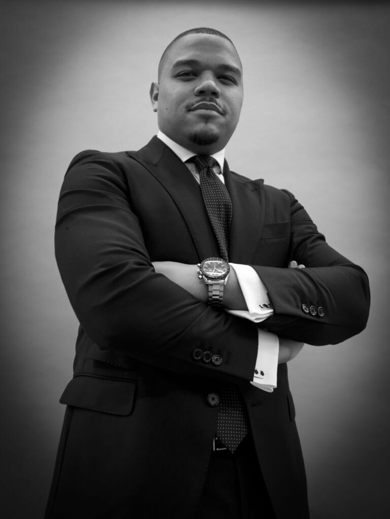 Dr. Kevin Murriel, pastor of Cascade United Methodist Church, in a black suit and polka dot tie, arms crossed, photographed in a confident black-and-white portrait