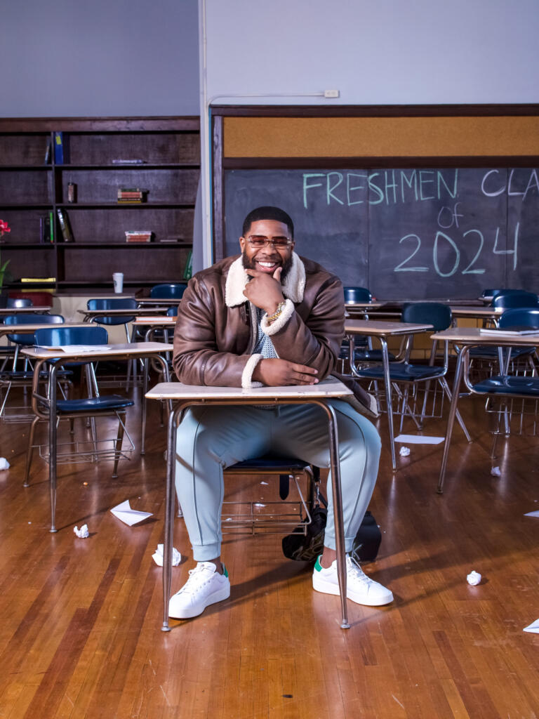 Yisrael Wright posing in a classroom desk for Hype Hair Magazine x Bronner Bros Freshman Class of 2024 issue, wearing a brown shearling jacket, light blue pants, and white sneakers