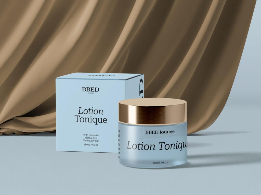 BBED Lounge Lotion Tonique jar and packaging box with gold accents displayed on a light blue surface in front of flowing beige fabric.